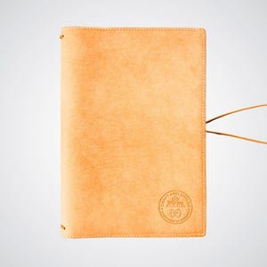 May include: A tan leather notebook cover with a brown leather strap and a circular logo that reads "Craft and Antics" with a small fox graphic inside the circle. The logo also says "Handmade in Canada".