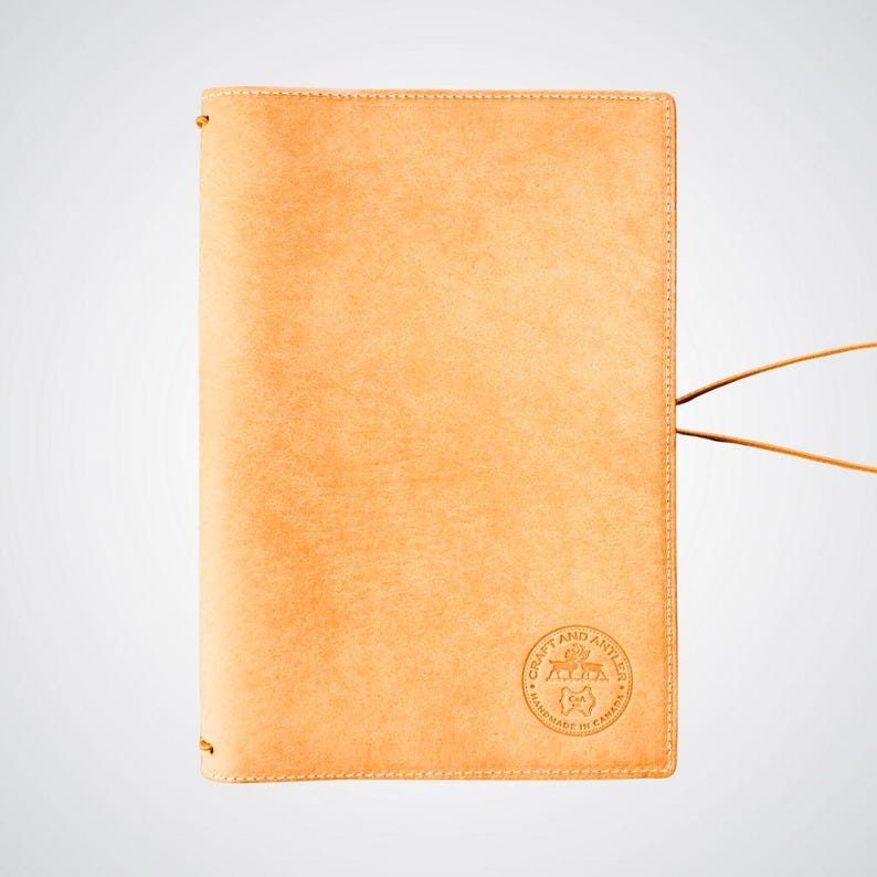 May include: A tan leather notebook cover with a brown leather strap and a circular logo that reads "Craft and Antics" with a small fox graphic inside the circle. The logo also says "Handmade in Canada".