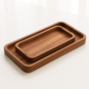 Pode incluir: Duas bandejas retangulares de madeira encaixadas. As bandejas são feitas de madeira de tons quentes com cantos arredondados. A bandeja maior tem aproximadamente 30 cm de comprimento, e a menor se encaixa dentro.