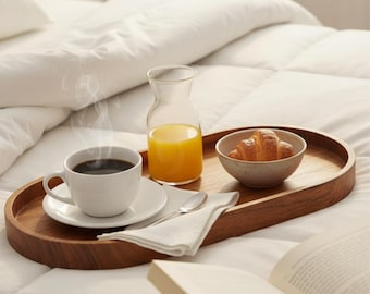 May include: A wooden serving tray holds a cup of coffee, a small bowl with a croissant, a glass carafe of orange juice, a spoon, and a folded napkin. The tray sits on a bed with white bedding. Steam rises from the coffee cup.