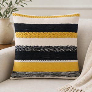 May include: A square throw pillow featuring horizontal stripes in cream, navy blue, and mustard yellow. The design includes textured, braided yarn accents in mustard yellow and navy blue, layered over a woven fabric base with a distinct pattern.