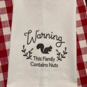 May include: White kitchen towel with black embroidery that reads "Warning This Family Contains Nuts" with a silhouette of a squirrel in the center.