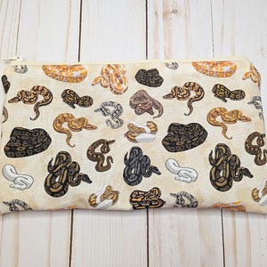 May include: A rectangular beige zipper pouch featuring a repeating pattern of colorful snakes in various poses. The snakes are in shades of brown, black, and white. The zipper is a light gold color.