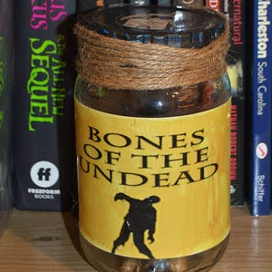May include: A glass jar with a brown twine tied around the lid. The jar has a yellow label with the text "Bones of the Undead" and a black silhouette of a zombie walking. The jar is sitting on a wooden surface.