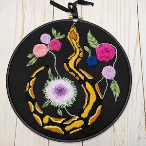 May include: Embroidered artwork on black fabric, featuring a yellow and brown snake intertwined with colorful flowers. The flowers include pink, purple, and blue accents, with green leaves. The artwork is framed in a black hoop and has a black ribbon for hanging.