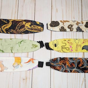 May include: Six fabric headbands with various patterns are displayed. The patterns include snakes, frogs, lizards, spiders, bats, and floral designs. Each headband has a black elastic band and black lace trim.