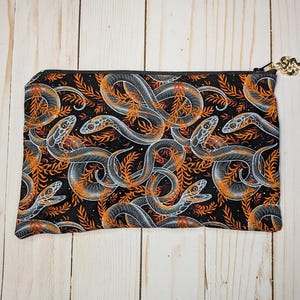 May include: Black zippered pouch featuring a repeating pattern of gray and orange snakes and foliage. The snakes have white highlights and are set against a black background. The zipper pull is gold-colored.