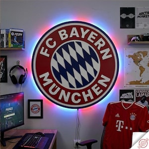 Pode incluir: Logotipo grande e iluminado do FC Bayern Munique com design vermelho e branco e padrão de losangos azuis e brancos. O logotipo é cercado por uma luz LED colorida. Uma camisa vermelha com o logotipo da equipe está em uma cadeira.