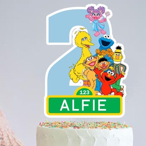 May include: A Sesame Street themed cake topper with a blue number 2, Big Bird, Elmo, Cookie Monster, and other characters. The topper also includes a green sign with the number 123 and the name Alfie.