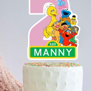 May include: A pink number two cake topper with Sesame Street characters including Big Bird, Elmo, Cookie Monster, Abby Cadabby, and Bert and Ernie. The topper has a green sign that says "123 MANNY".