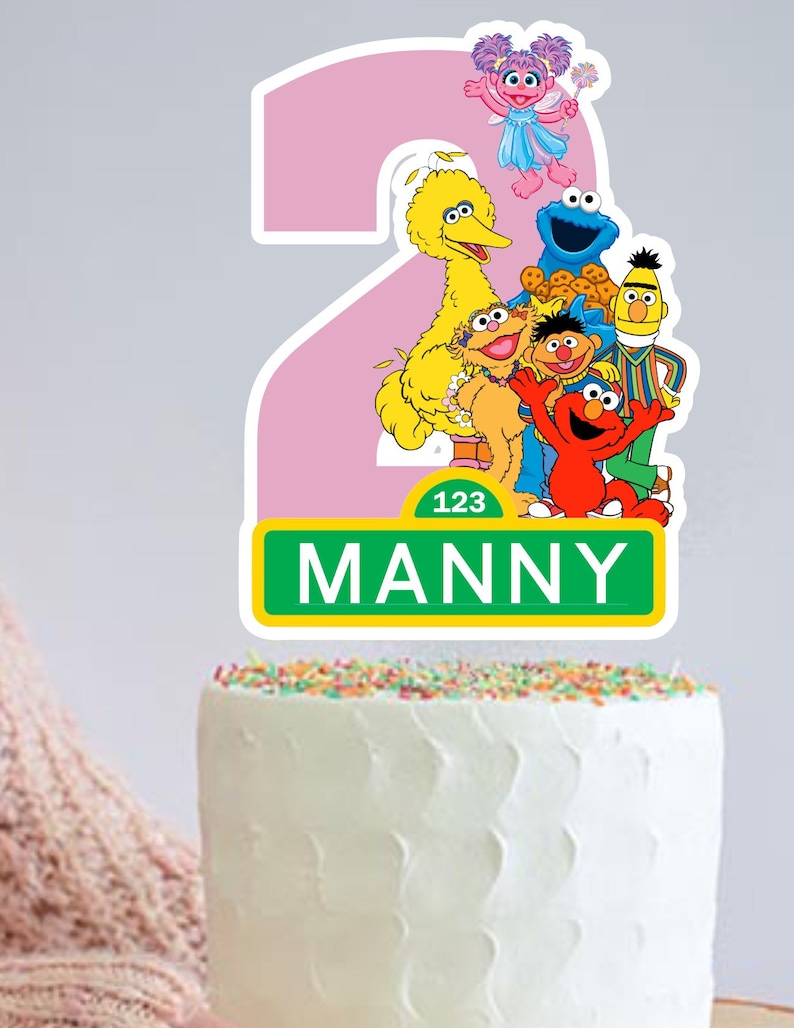May include: A pink number two cake topper with Sesame Street characters including Big Bird, Elmo, Cookie Monster, Abby Cadabby, and Bert and Ernie. The topper has a green sign that says "123 MANNY".