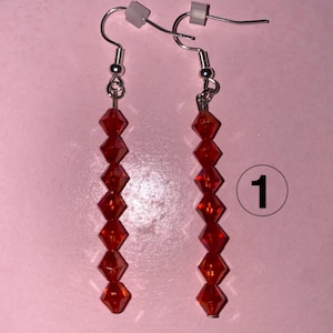 May include: A pair of red beaded dangle earrings. Each earring has a series of faceted, red beads strung on silver-coloured wire. The earrings have fish hook style ear wires. The background is pink.