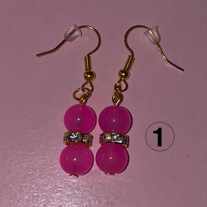 Op de afbeelding: Een paar oorhangers met goudkleurige haken. Elke oorbel heeft twee helder roze, ronde kralen gescheiden door een goudkleurige band met heldere strass-steentjes. De achtergrond is effen roze.