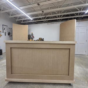 May include: A large, unfinished light brown wooden bar counter featuring a rectangular front panel with vertical slats. The structure includes two matching curved side panels with vertical fluting, all mounted on small wheels for mobility.