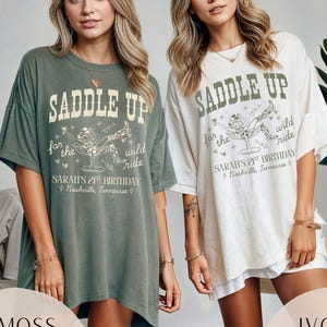 May include: Two women wearing oversized t-shirts with a graphic print of a woman riding a horse with the text "Saddle Up for the Wild Ride" and "Sarah's 21st Birthday Nashville, Tennessee". The t-shirt on the left is moss green and the t-shirt on the right is ivory.
