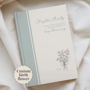 May include: A hardcover journal with a cream-colored cover featuring a sage green vertical stripe on the left. The text reads Chapter Thirty, EVERY NEW CHAPTER BRINGS GROWTH, and keep blooming. A floral bouquet illustration and the initials L.M. appear.