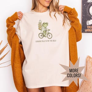 May include: Cream-colored t-shirt featuring a whimsical illustration of a frog riding a bicycle, wearing glasses and a lab coat, carrying a stack of items. The text "SLINGING PILLS TO PAY THE BILLS" is printed below the graphic. A starburst graphic says "MORE COLORS!"