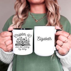 May include: White ceramic mug with a black rim and handle. The mug has black text that reads "Chapter 40 and still writing my story" with a floral design. The mug is next to a second white ceramic mug with a black rim and handle. The second mug has black text that reads "Elizabeth".