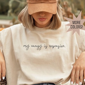 May include: Beige crew neck t-shirt with the phrase "my energy is expensive" in black script. The shirt is worn by a person wearing a brown corduroy baseball cap. A starburst graphic indicates more colors are available.