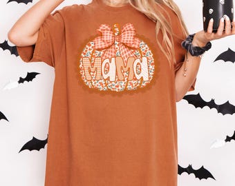 May include: A burnt orange t-shirt featuring a pumpkin design with the word "MAMA" in a decorative font. The pumpkin has a floral and plaid pattern with a matching bow. The shirt is a comfortable, casual style.