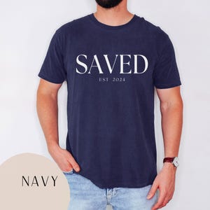 May include: A navy blue t-shirt with the word "SAVED" in white lettering and the text "EST 2024" underneath.