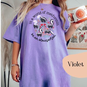 K&ouml;nnte beinhalten: Ein lilafarbenes Oversize-T-Shirt mit einer Grafik von Stinktieren und dem Text "In einer Welt von Metformin sei ein Olmesartan".