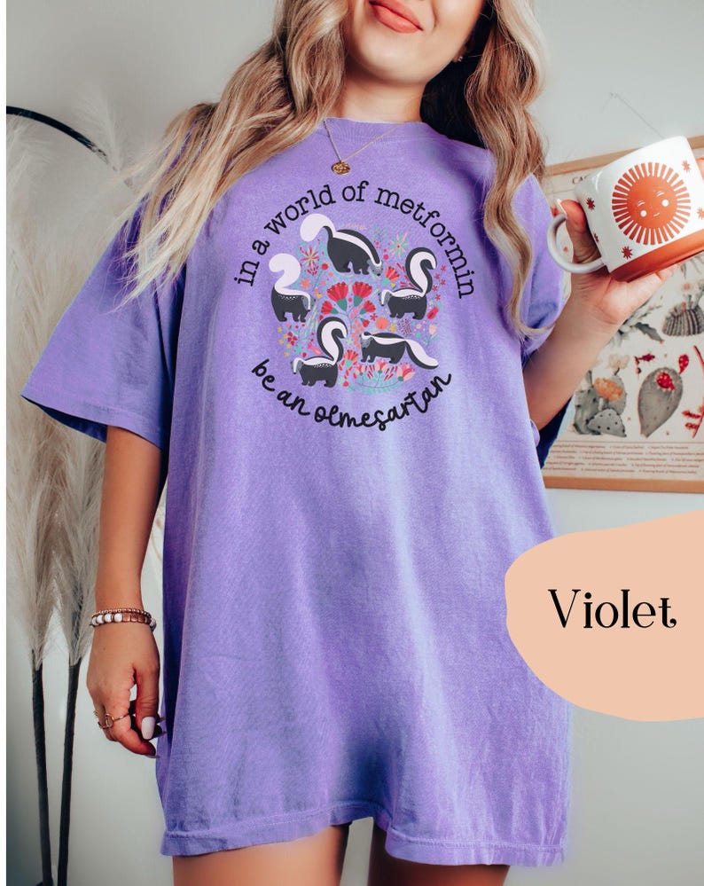 K&ouml;nnte beinhalten: Ein lilafarbenes Oversize-T-Shirt mit einer Grafik von Stinktieren und dem Text "In einer Welt von Metformin sei ein Olmesartan".