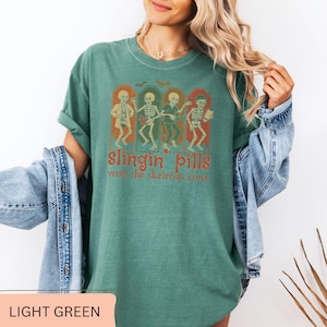 May include: Light green t-shirt with the text "Slingin' Pills with the skeleton crew." The graphic features four skeletons in orange frames. The shirt is a relaxed fit and has rolled-up sleeves.