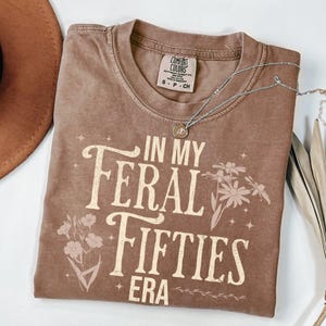 May include: A brown Comfort Colors t-shirt with the text "IN MY FERAL FIFTIES ERA" in a vintage font. The shirt features floral graphics and a silver necklace with a pendant. The shirt is a small size.