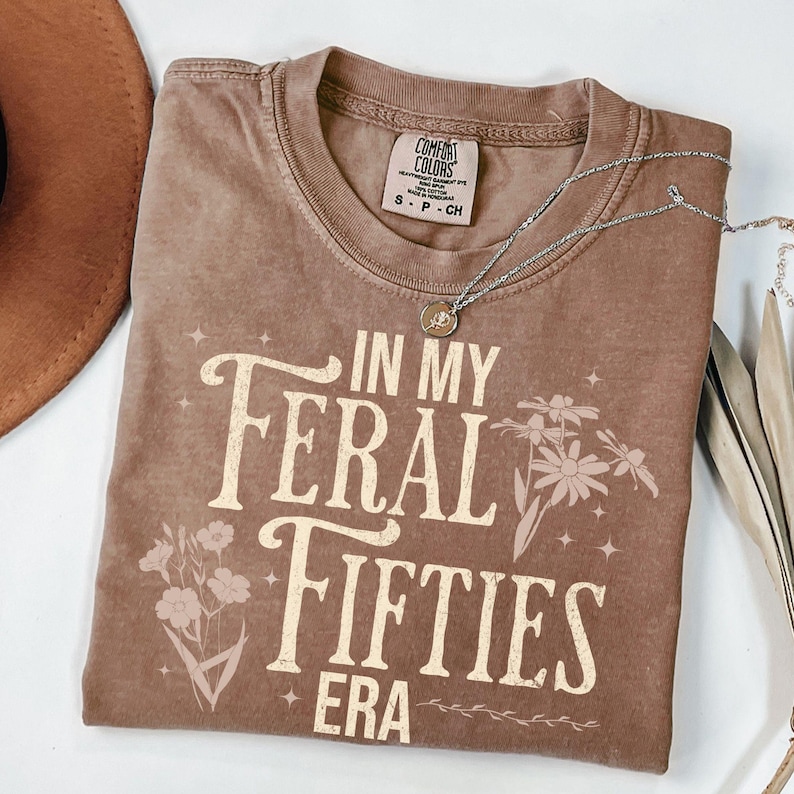 May include: A brown Comfort Colors t-shirt with the text "IN MY FERAL FIFTIES ERA" in a vintage font. The shirt features floral graphics and a silver necklace with a pendant. The shirt is a small size.