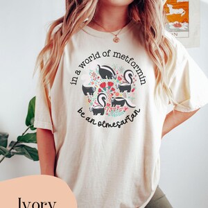 K&ouml;nnte beinhalten: Ein T-Shirt in Elfenbein mit einem Motiv aus Stinktieren und Blumen. Der Text auf dem T-Shirt lautet "In einer Welt von Metformin, sei ein Olmesartan."