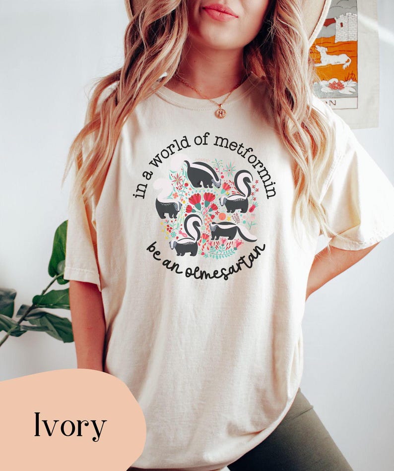 K&ouml;nnte beinhalten: Ein T-Shirt in Elfenbein mit einem Motiv aus Stinktieren und Blumen. Der Text auf dem T-Shirt lautet "In einer Welt von Metformin, sei ein Olmesartan."