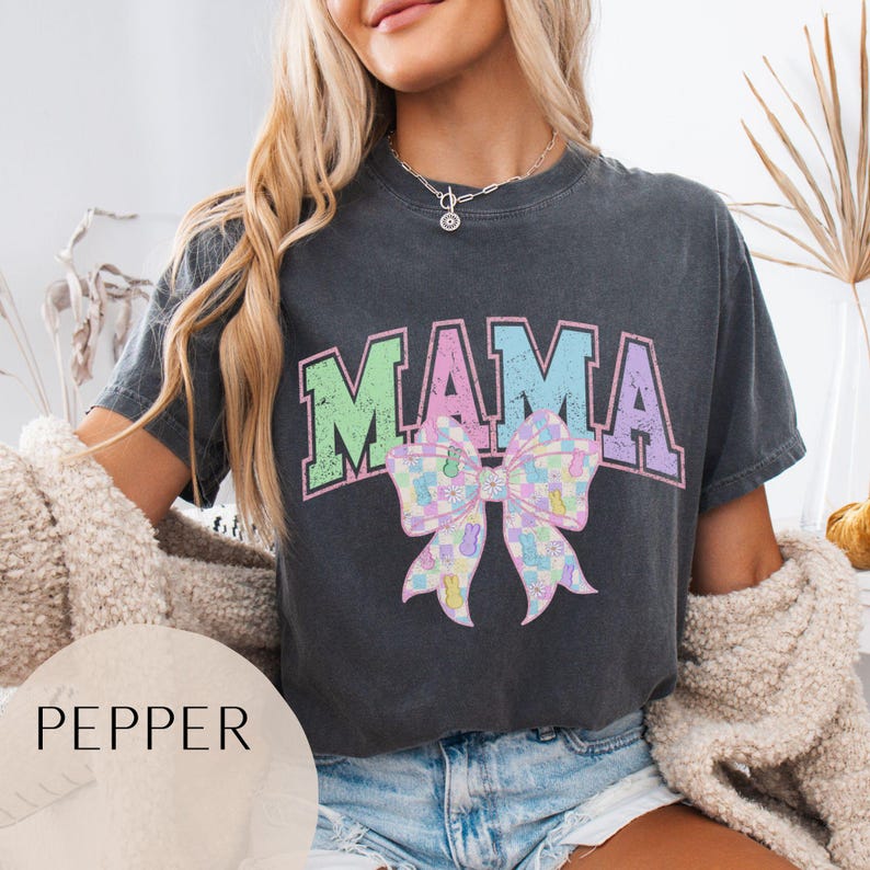May include: A dark gray t-shirt with the word "MAMA" printed in pastel green, pink, and purple letters. A pastel checkered bow with pastel bunnies is below the word "MAMA".