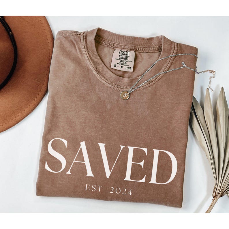 May include: A brown t-shirt with the word "SAVED" in white text and the words "EST 2024" below it.