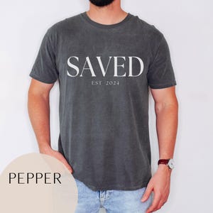 May include: A dark gray t-shirt with the word "SAVED" printed in white on the front. The text "EST 2024" is printed below the word "SAVED".