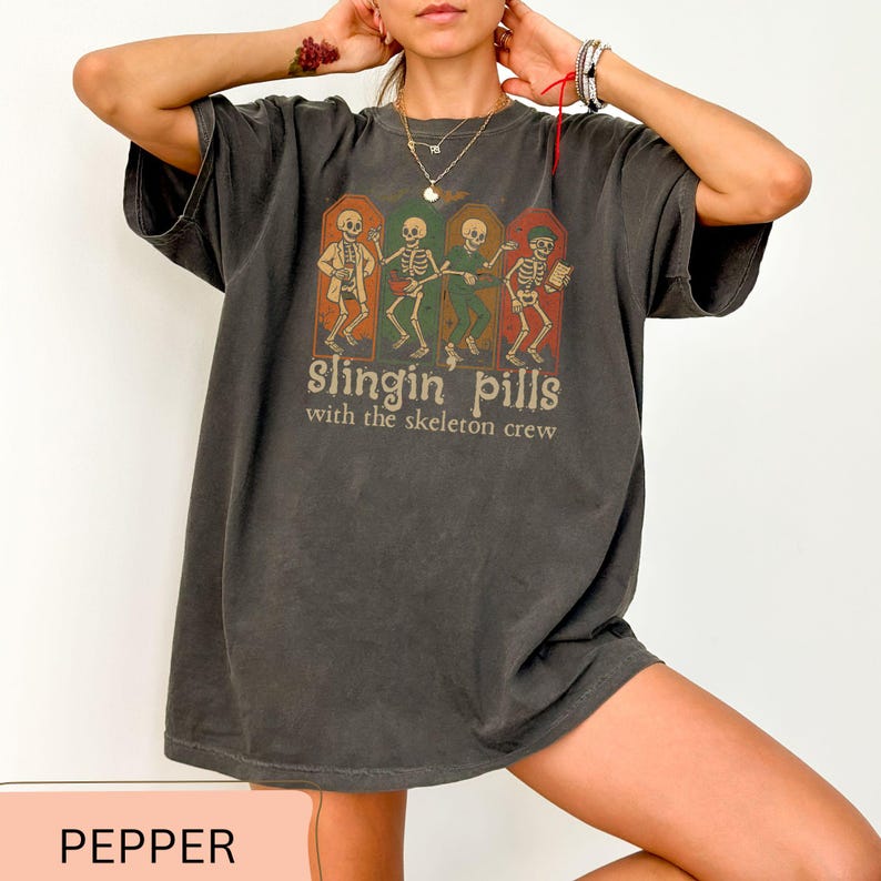 May include: Dark grey t-shirt with a graphic of skeletons in frames, and the text "slingin' pills with the skeleton crew." The design has a vintage, slightly distressed look. The t-shirt is pepper-colored.