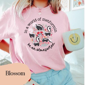 K&ouml;nnte beinhalten: Ein pinkfarbenes T-Shirt mit einem Motiv aus Stinktieren und Blumen. Der Text auf dem Shirt lautet "in a world of metformin be an olmesartan".