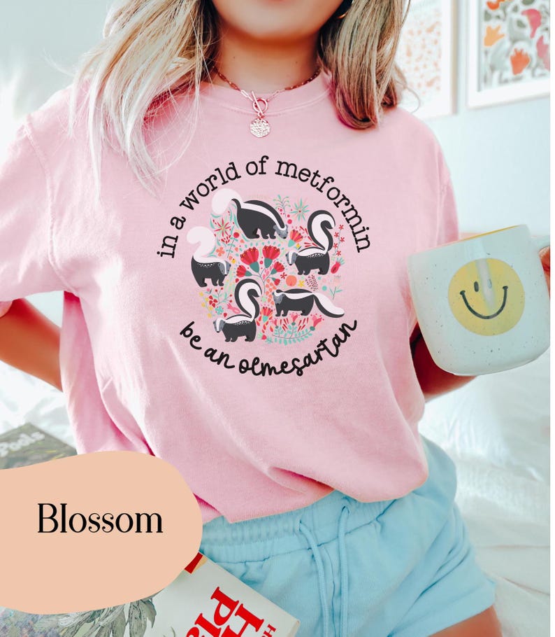 K&ouml;nnte beinhalten: Ein pinkfarbenes T-Shirt mit einem Motiv aus Stinktieren und Blumen. Der Text auf dem Shirt lautet "in a world of metformin be an olmesartan".