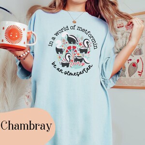 K&ouml;nnte beinhalten: Ein hellblaues T-Shirt mit einer Grafik von Stinktieren und Blumen. Der Text auf dem Shirt lautet "In einer Welt von Metformin sei ein Olmesartan".