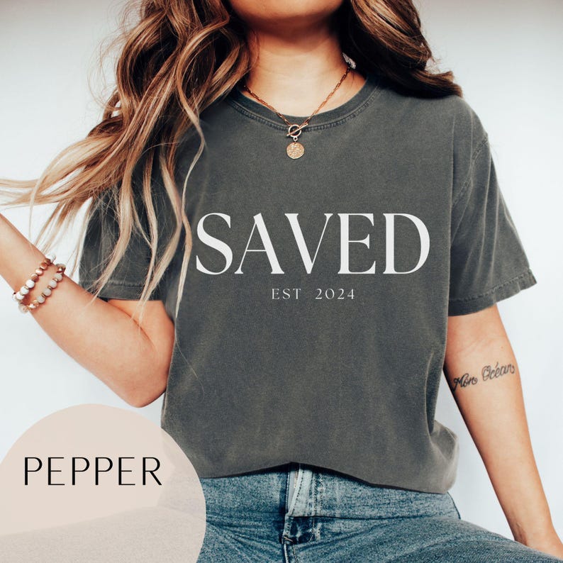 May include: A gray t-shirt with the word "SAVED" printed in white, with the text "EST 2024" underneath. The shirt is being worn by a person with long brown hair.