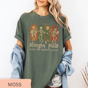 May include: Moss green t-shirt with a graphic of four dancing skeletons in arched frames. The text "slingin pills with the skeleton crew" is below the skeletons. The shirt has a relaxed fit.