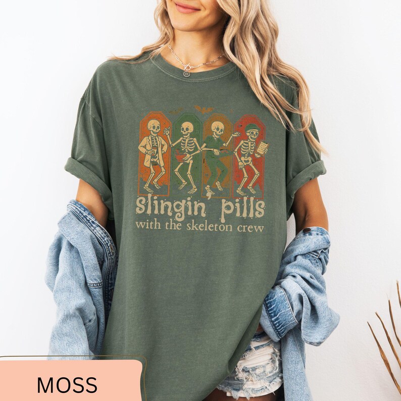 May include: Moss green t-shirt with a graphic of four dancing skeletons in arched frames. The text "slingin pills with the skeleton crew" is below the skeletons. The shirt has a relaxed fit.