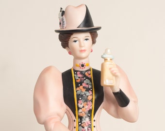 May include: A porcelain figurine of a woman wearing a pink dress with black and floral accents, a black and white hat, and holding a bottle of perfume.