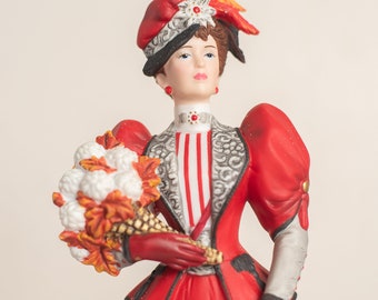 May include: A porcelain figurine of a woman in a red dress and a black and white hat holding a bouquet of white flowers and a corn stalk.