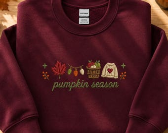 Può includere: Una felpa girocollo bordeaux con un ricamo raffigurante una foglia d'acero rossa, una catena di luci, un cesto di mele e un maglione color crema con un cuore rosso. Il testo "pumpkin season" è ricamato in verde.