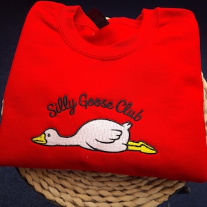 Può includere: Una felpa rosso brillante con la scritta ricamata "Silly Goose Club" sopra un'oca dei cartoni animati. L'oca è bianca con becco e zampe gialle. La felpa è piegata su una superficie tessuta.