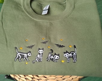 Puede incluir: Una sudadera verde con un diseño bordado de cuatro gatos esqueleto y murciélagos. Los gatos son negros y blancos con estrellas amarillas a su alrededor.