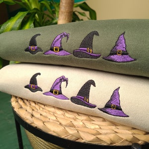 Può includere: Due felpe con cappelli da strega ricamati. La felpa superiore è verde oliva e quella inferiore è beige. Ogni felpa ha cinque cappelli da strega ricamati. I cappelli sono viola con bordi neri e fibbie gialle.