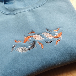 May include: A light blue sweatshirt with an embroidered design of orange and black fish swimming in a school.