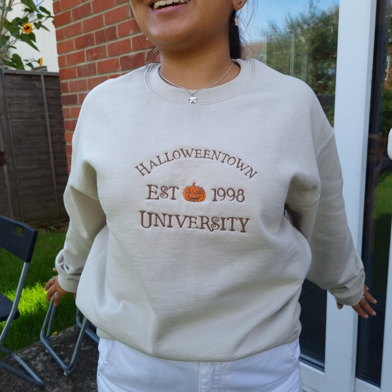 May include: A beige sweatshirt with the text "HALLOWEENTOWN EST 1998 UNIVERSITY" embroidered on the front. The text is in brown and features a small orange pumpkin with a black face.